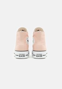 Zapatillas de lona altas de color rosa con suelas de goma blancas, detalles en negro y un parche con un logo circular en la parte trasera. Textura suave y diseño sencillo.