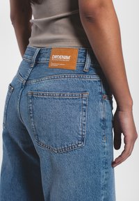 Jeans in denim blu con vita alta, dotati di due tasche posteriori e di una toppa in pelle marrone chiaro con testo bianco sulla cintura.