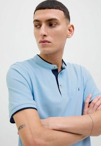 Polo shirt azzurro chiaro con colletto, caratterizzato da dettagli blu scuro e due bottoni. Tessuto testurizzato con maniche corte, branding minimo visibile.