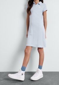 Meisje in een lichtblauwe polojurk, blauwe sokken en witte hoge Converse sneakers, staande tegen een witte muur en een grijze vloer.