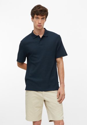 Junger Mann, der nach vorne blickt und ein dunkelblaues kurzärmeliges Poloshirt sowie hellbeige knielange Shorts vor einem weißen Hintergrund trägt.