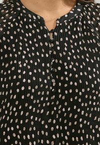 Svart, plisserad blus med vita och beigea prickar. Halsringning med nyckelhål och knappar. Något textilaktig, avslappnad passform.