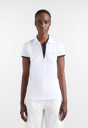 Giovane donna con capelli scuri intrecciati che indossa una polo bianca a maniche corte con bordatura blu navy e pantaloni bianchi davanti a uno sfondo semplice.