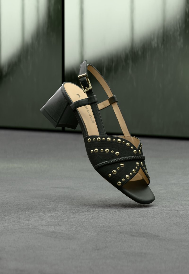 Μαύρο σαμπαντό με ανοιχτά δάχτυλα, τακούνι τύπου block, χρυσά τρουκς, λεπτομέρεια πλεγμένου λουριού και λουράκι με αγκράφα στον αστράγαλο, πάνω σε γκρι υφή.