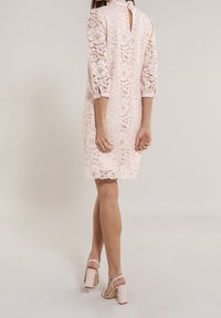 Šifónové šaty v jemně růžové, tříčtvrteční rukávy, zdobený lem, vysoký výstřih s klíčovou dírkou na zádech, doplněné světle zbarvenými sandály na blokovém podpatku.