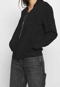 Black bomber jacket with a zip front, round neckline, and side pockets, featuring a smooth texture and fitted cuffs. Paired with dark jeans.