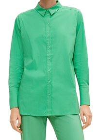 Camicia verde con bottoni, realizzata in tessuto morbido, con colletto classico, maniche lunghe e orlo dritto, indossata con pantaloni abbinati.