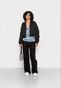 Mulher com cabelo encaracolado vestindo uma jaqueta bomber preta, blusa azul com padrão floral, calças largas pretas, sapatos brancos, óculos de sol e segurando uma bolsa com estampa de zebra.