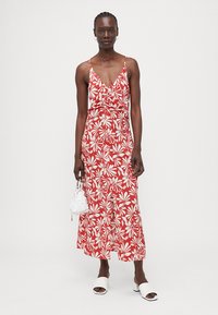 Rotes Maxi-Kleid mit floralen Mustern in Weiß, V-Ausschnitt, verstellbaren Trägern und einer Schleife in der Taille. Ein Paar weiße Sandalen und eine Tasche.