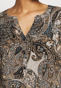 Schwarz, beige und blaue gemusterte Bluse mit V-Ausschnitt und Knopfverschluss. Merkmale sind aufwendige Paisley- und Blumendesigns auf einem leichten Stoff.