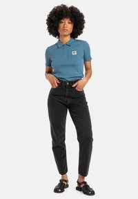 Blue polo shirt with short sleeves and a collar, paired with high-waisted black jeans and black sandals. Features a small logo patch on the shirt.
