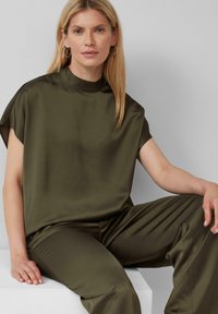 Blouse satin vert olive avec un col montant et des manches courtes, associée à un pantalon satin assorti. Texture lisse, coupe décontractée.