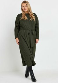 Robe en tricot côtelé vert olive, à manches longues et avec une ceinture à nouer à la taille. La robe a une coupe décontractée et une longueur midi. Bottines noires.