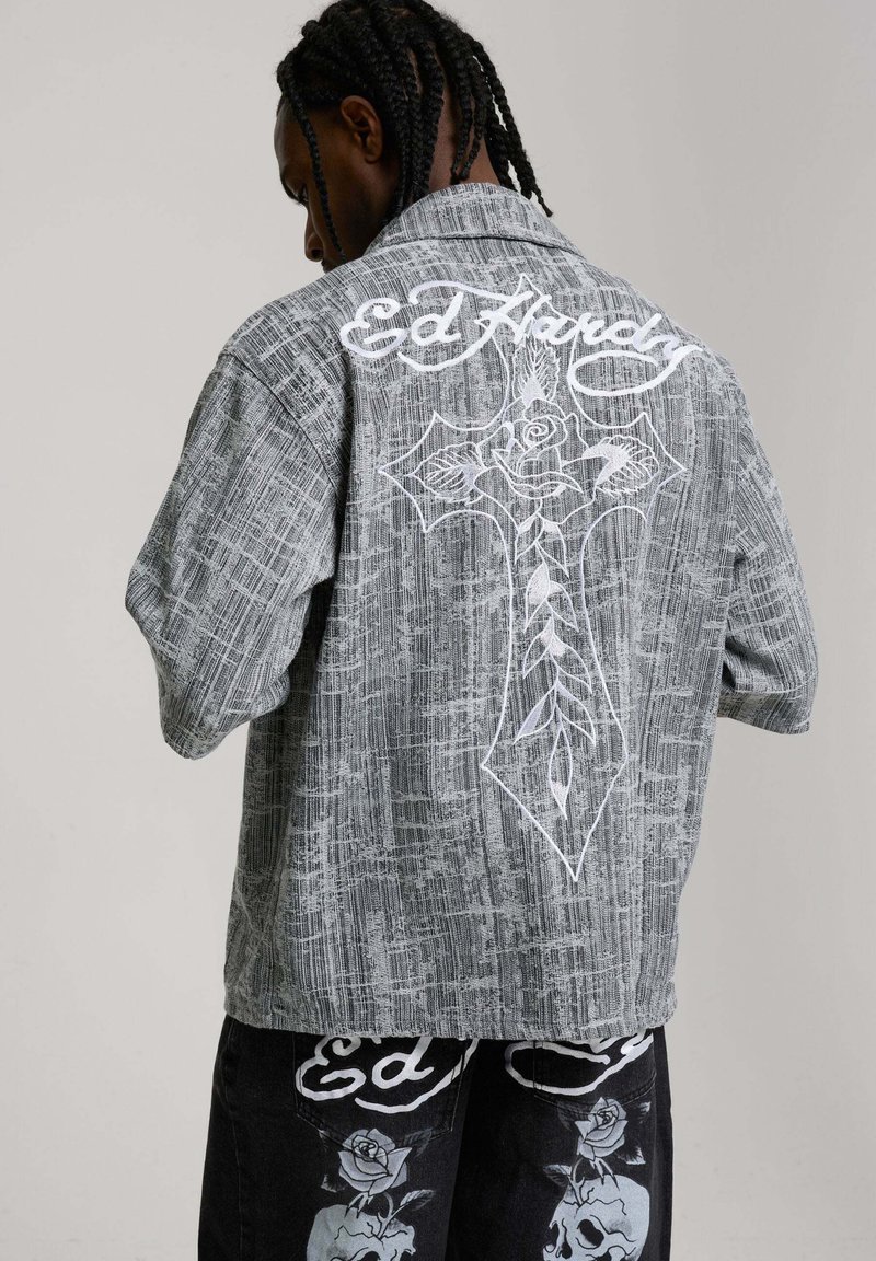 Mann mit geflochtenem Haar trägt eine graue texturierte Jacke mit großem weißem Kreuz und Rosendesign sowie dem Schriftzug „Ed Hardy“ auf dem Rücken, kombiniert mit schwarzen Hosen.