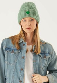 Groen geribbelde brei-beret met een klein geborduurd hartlogo, gedragen met een spijkerjack over een wit shirt. Zachte textuur en aansluitend ontwerp.