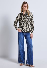 Leopardenmuster-Hemd in Beige und Schwarz, kombiniert mit weit geschnittenen blauen Jeans und beigen Sandalen, vor einem neutralen Hintergrund.