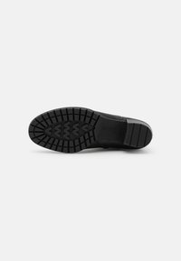 Suela de zapato negra con patrones de tracción profundos, tacón rectangular y nombre de la marca grabado cerca del arco, mostrado sobre un fondo blanco.
