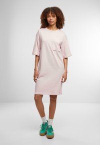 Rochie T-shirt oversized roz deschis, cu mâneci scurte, guler rotund, buzunar frontal și tiv drept. Asortată cu sneakers verzi și șosete albe.