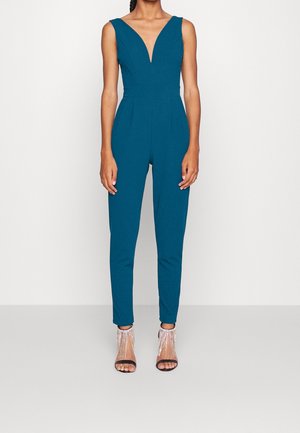 Teal jumpsuit dengan leher V yang dalam, bodice yang pas, dan kaki yang menyempit. Tekstur halus, desain tanpa lengan, dipadukan dengan sandal hak tinggi yang bertali.