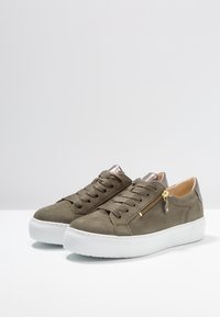 Zielone zamszowe sneakersy w odcieniu oliwkowym z białą podeszwą, z metalowym zamkiem po boku, dopasowanymi sznurowadłami oraz metalicznym akcentem na pięcie.