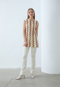 Beige, ermeløs tunika med zigzag-mønster i brune toner, kombinert med hvite, tettsittende bukser og netting ankelsko med spisse tær.