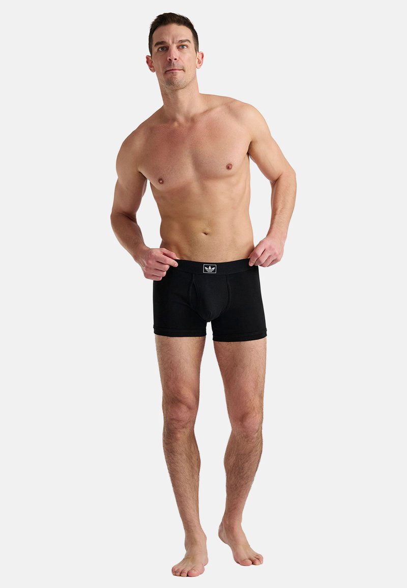 Homme torse nu portant un boxer Adidas noir, debout pieds nus sur un fond gris clair uni, ajustant la ceinture avec ses mains.