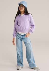 Lila tröja med lång ärm och krage, kombinerad med en vit skjorta med volanger, ljusblå vida jeans och grå sneakers. Ljusblå keps.