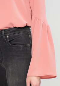 Blouse rose en tissu texturé avec manches évasées, associée à un jean en denim noir. Mise au point sur le détail de la manche et la couleur du vêtement.