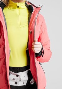 Personne portant un haut zippé jaune vif sous une veste matelassée corail ouverte, avec un pantalon de neige noir et blanc visible à la taille.