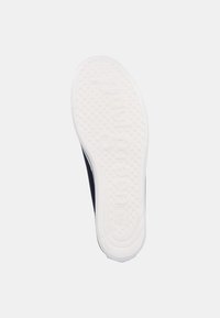 Suela de goma blanca con un patrón texturizado, con puntos elevados y letras en relieve, unida a la parte superior del zapato de color azul marino oscuro.