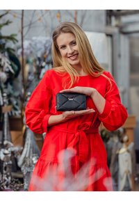 Black quilted wallet with a zip closure and logo accent, held by a person in a red dress. Textured material, compact rectangular shape.
