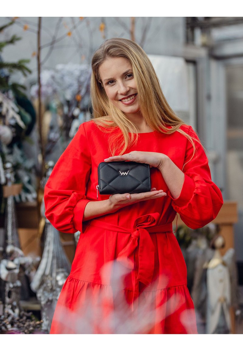 Black quilted wallet with a zip closure and logo accent, held by a person in a red dress. Textured material, compact rectangular shape.