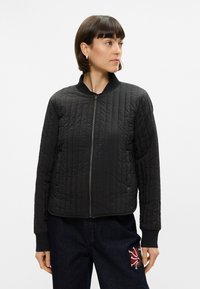 Veste bomber matelassée noire avec col côtelé, fermeture éclair et tissu texturé. Dotée de poignets ajustés et d'une doublure lisse.