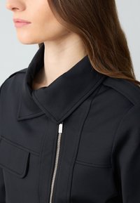 Zwarte jas met hoge kraag, ritsluiting en een voorzak. Het materiaal heeft een gladde textuur met minimale stikdetails.