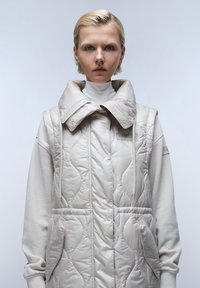 Gilet beige imbottito con ampio colletto e zip, caratterizzato da motivi trapuntati, tasche frontali e una texture morbida, indossato sopra un dolcevita.