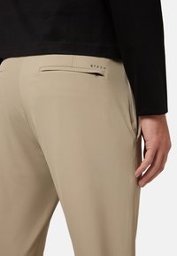 Pantalones beige confeccionados en tela suave que presentan un bolsillo trasero con cremallera. La cinturilla es sencilla y el material tiene un acabado mate.