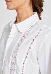 Camicia bianca con bottoni, caratterizzata da dettagli in pizzo lungo le cuciture verticali, con colletto classico e texture liscia.