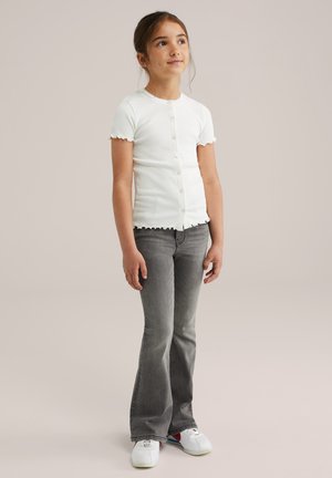 Jong meisje staat, gekleed in een witte knoopblouse met korte mouwen, grijze flare jeans en witte sneakers, terwijl ze iets omhoog en rechts kijkt.