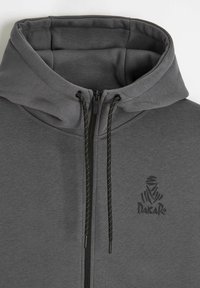Ciemnoszary hoodie na zamek z czarnymi sznurkami, wyściełanym kołnierzem oraz małym logo Dakar na lewej piersi.