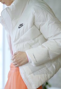 Balta pūsta Nike striukė su aukštu apykakle, su juodu logotipu ant krūtinės ir detale "THERMA-FIT" ant rankovės. Lygus, blizgus paviršius.
