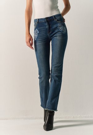 Mujer con vaqueros vaqueros azules con bordado floral cerca de los bolsillos, bajos deshilachados y botas negras de tacón hasta el tobillo, apoyada contra una pared lisa.