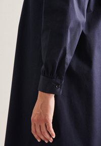 Tmavě modrá látková košile s dlouhými rukávy. Rukáv je nažehlený a má malý knoflík pro zavírání, což dodává designu jemný detail.