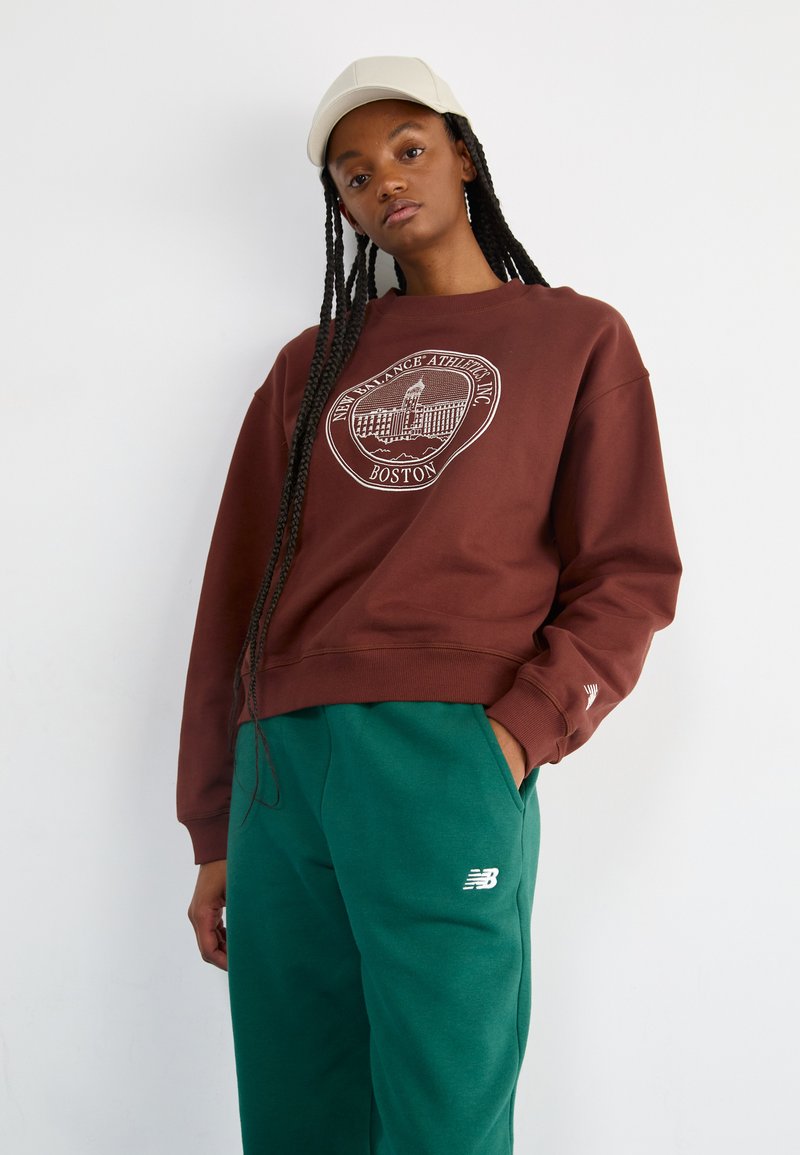 Bruine sweatshirt met een wit Boston-logo, gecombineerd met groene joggingbroeken met een klein wit New Balance-logo op het been.