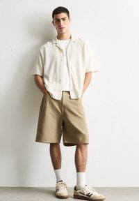Light beige short-sleeve button-up shirt over a white t-shirt, paired with khaki shorts and cream sneakers with brown accents.
