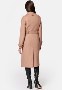 Beżowy trencz z wełny z paskiem w talii, długimi rękawami i rozcięciem z tyłu. Posiada klasyczny kołnierz i zapinane mankiety.