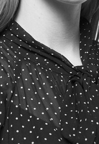 Černá, průhledná blůza s bílými puntíky, se zavázaným výstřihem a jemným drapováním. Materiál vypadá lehký a vzdušný.