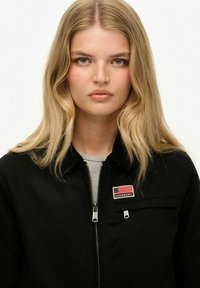 Zwarte zip-up jas met een borstzak met een klein label en een vlag ontwerp. Eenvoudig ontwerp met een gladde textuur.