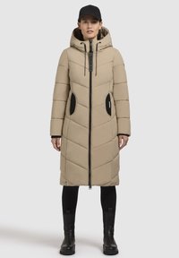 Beige lang pufferjakke med hætte, quilts med diamantmønster, sorte lynlås-lommer og lynlåslukning foran. Båret over sorte leggings og støvler.