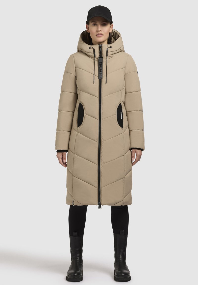 Beige lang pufferjakke med hætte, quilts med diamantmønster, sorte lynlås-lommer og lynlåslukning foran. Båret over sorte leggings og støvler.