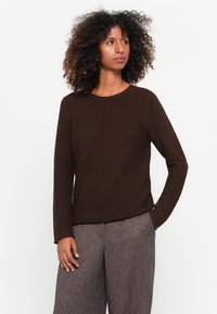 Brauner gerippter Pullover mit rundem Ausschnitt und langen Ärmeln, kombiniert mit strukturierten, hellgrauen, weit geschnittenen Hosen. Glatte Stofftextur.